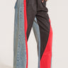 A person is posing in the HARDEST TO LOVE COLORBLOCK DENIM PANT, which are stylish, high-rise, wide-leg pants featuring a unique color-block design. The pants showcase denim panels along the sides, black sections in the middle, and red sections on the front. The person is accessorizing with gold bracelets and wearing black and white shoes.