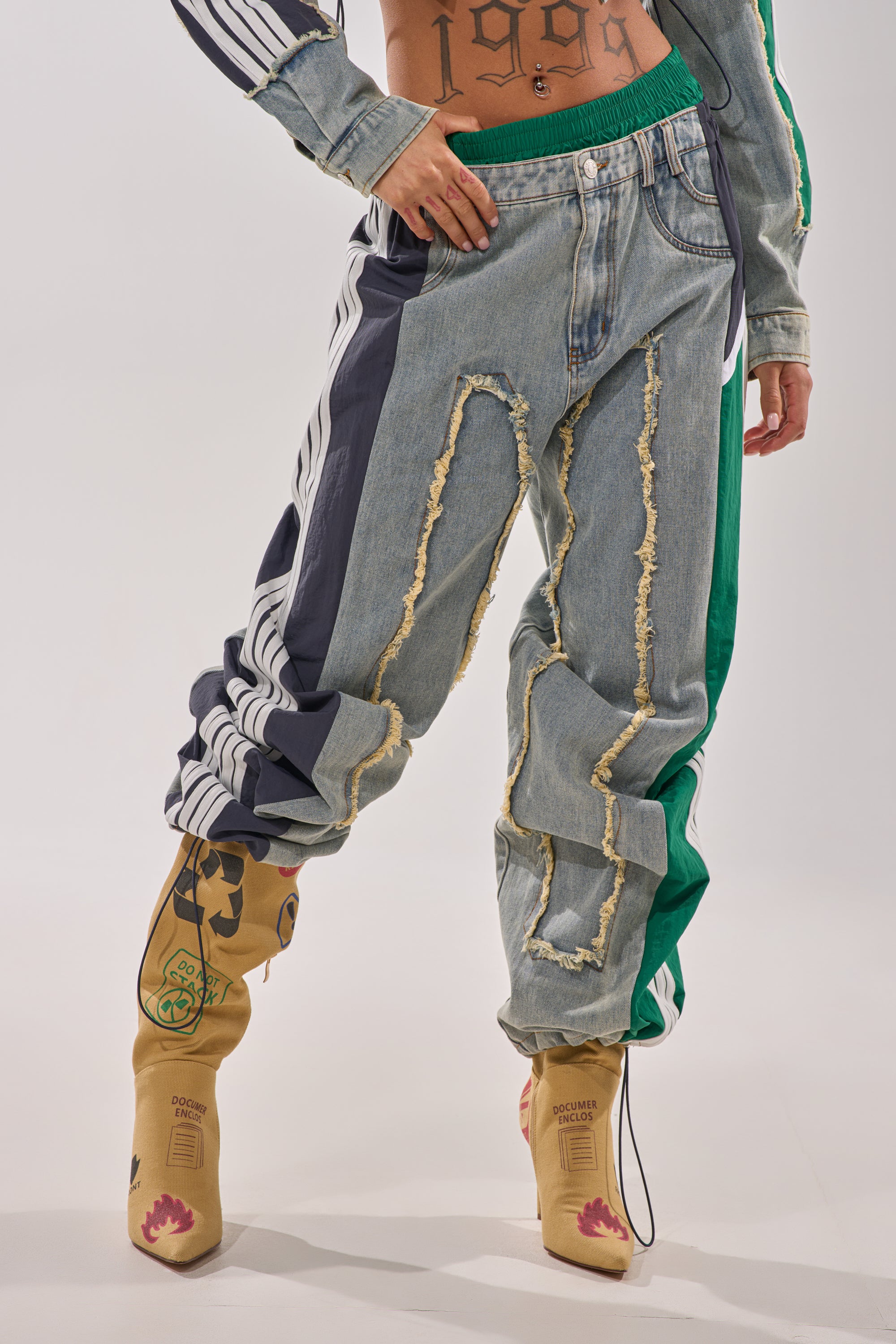 A person wears the ALL AROUND THE WORLD PATCHWORK DENIM JOGGER PANT IN GREEN MULTI with frayed seams, patterned side stripes, and tan artistic boots, standing against a plain background; only the lower torso and legs are shown.