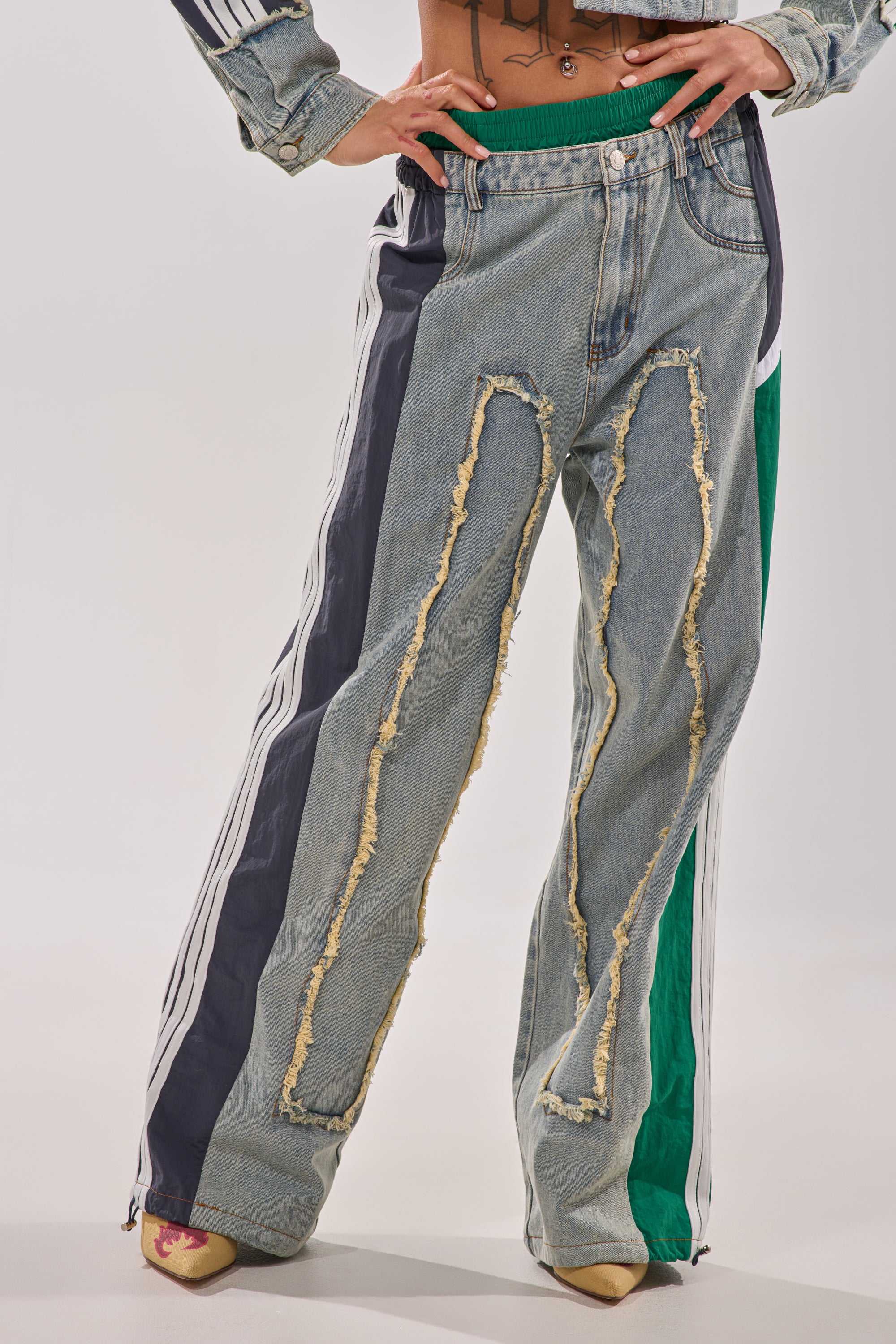 A person wears the ALL AROUND THE WORLD PATCHWORK DENIM JOGGER PANT IN GREEN MULTI, featuring ripped panels, green and black sporty side stripes, and pointed tan heels. Only the lower torso and legs are visible.
