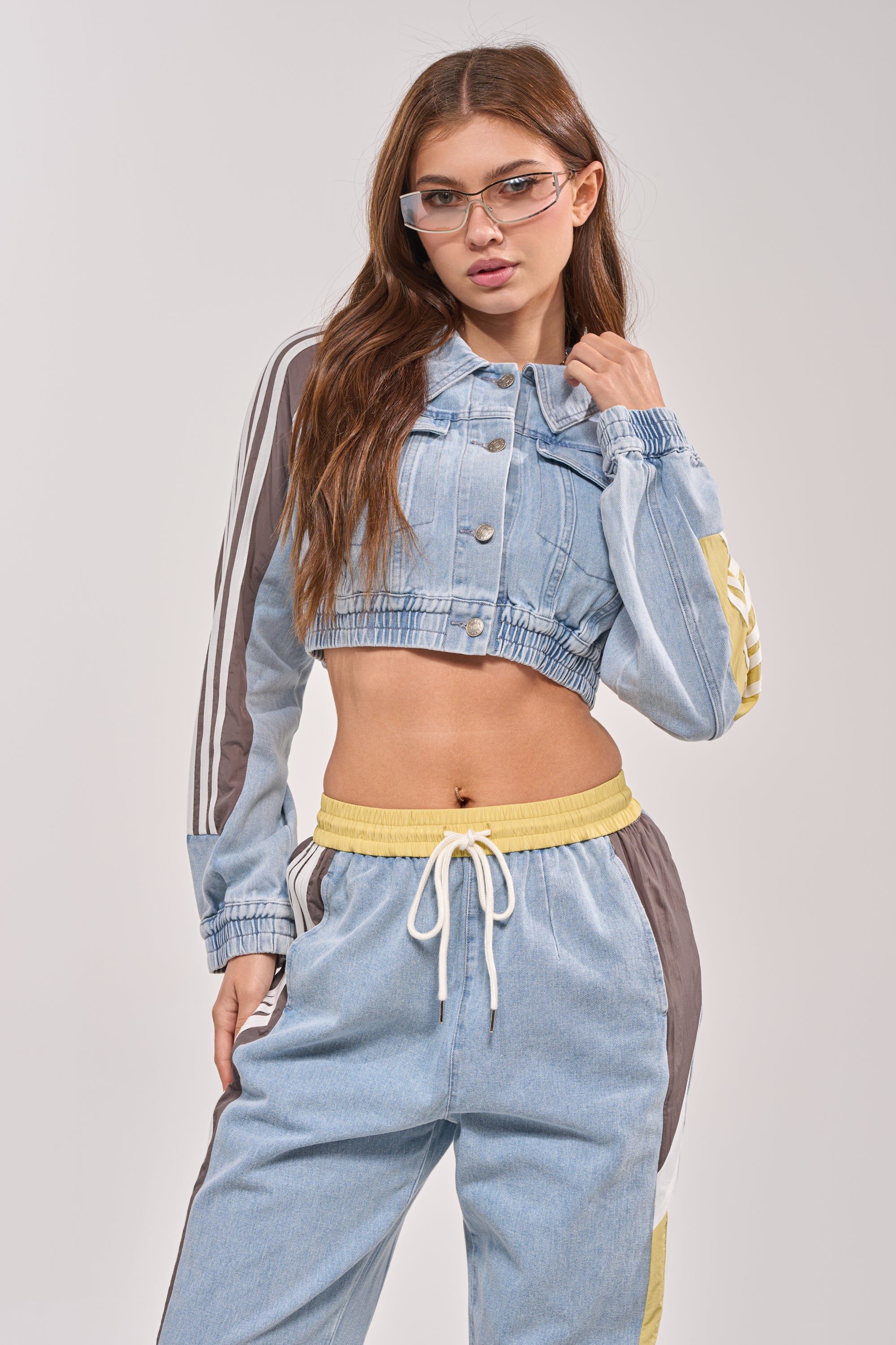 A woman with long hair and glasses showcases the NEVER BETTER DENIM JACKET, featuring striped sleeves, paired with matching drawstring track pants. She stands confidently, one hand on her hip, embodying a retro athleisure vibe. 3