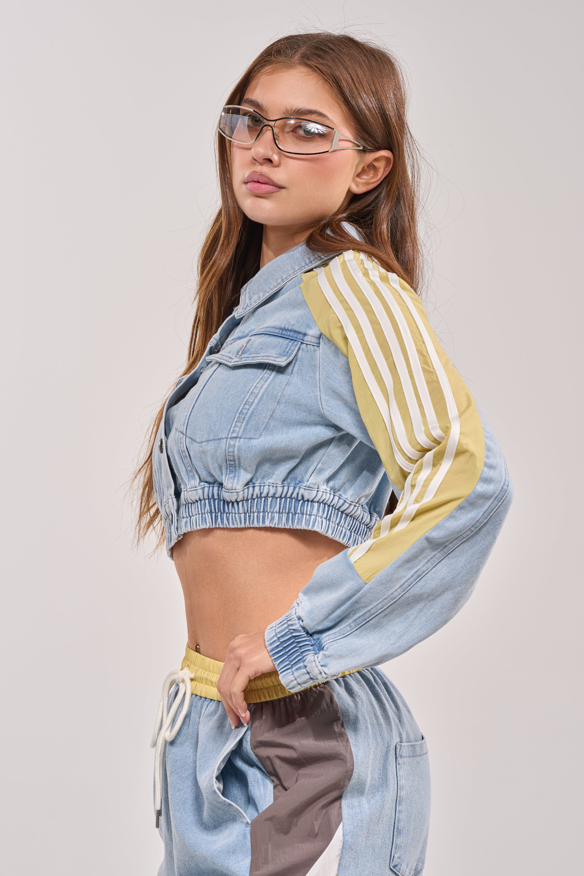 A young woman with long brown hair and clear glasses stands in profile against a plain background, wearing the NEVER BETTER DENIM JACKET with yellow and white striped sleeves and matching blue jogger pants with white drawstrings. 4