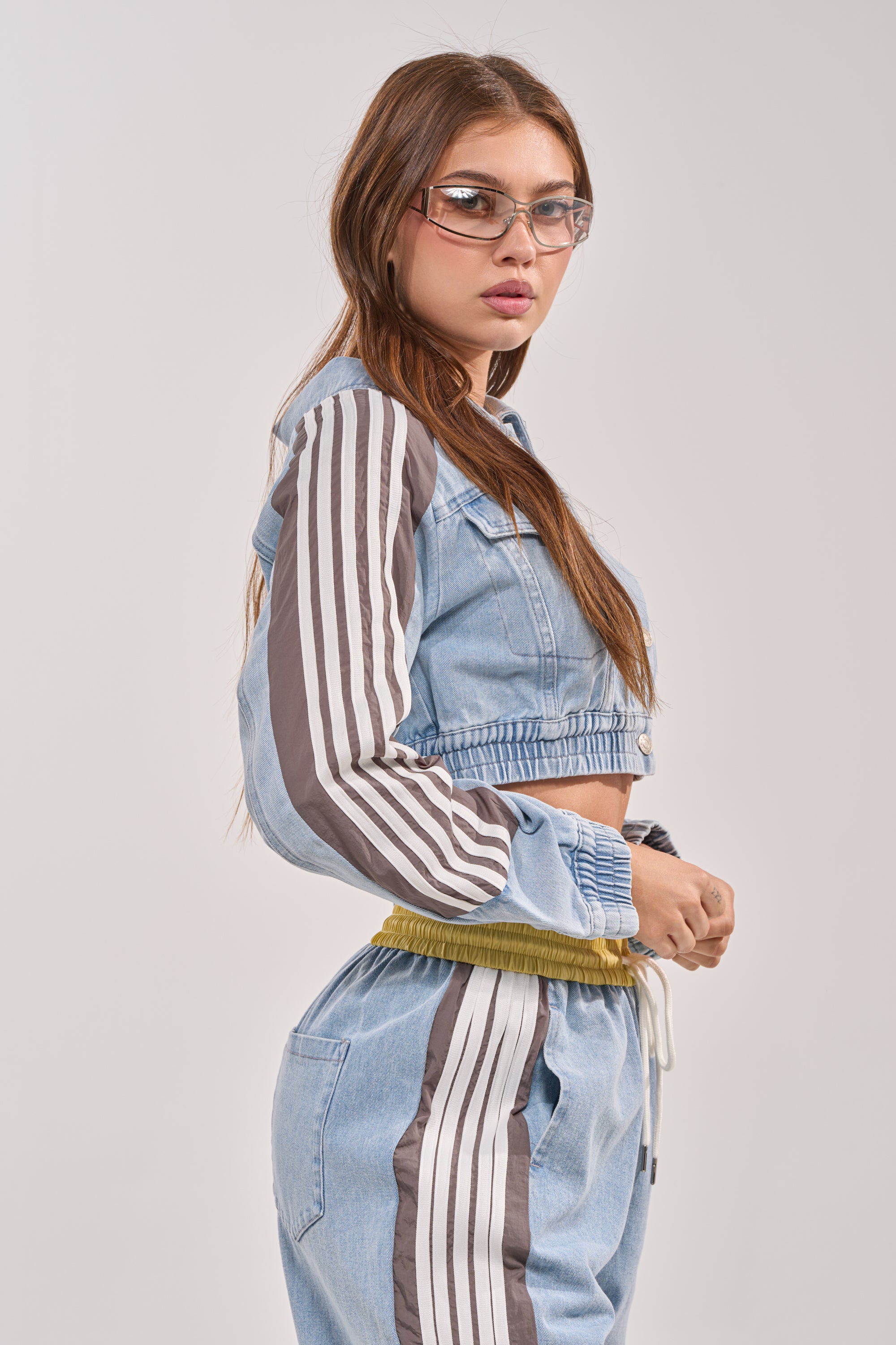 A woman with long brown hair and clear glasses wears the NEVER BETTER DENIM JACKET with striped sleeves and matching high-waisted joggers, standing sideways against a plain background. 5