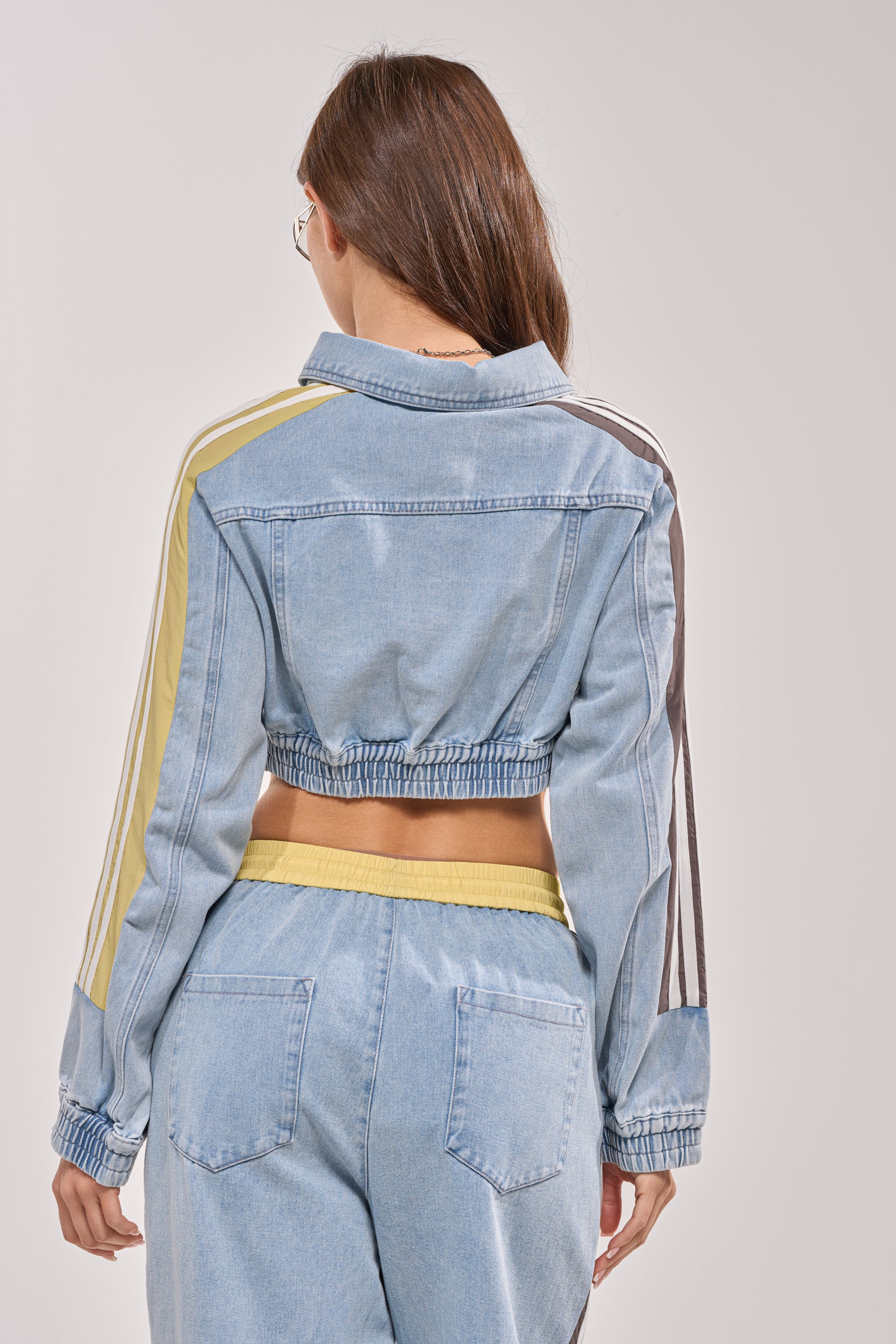 A woman with long brown hair is seen from behind wearing the NEVER BETTER DENIM JACKET, a light blue cropped denim jacket with striped sleeve detailing, paired with matching high-waisted denim pants. 6