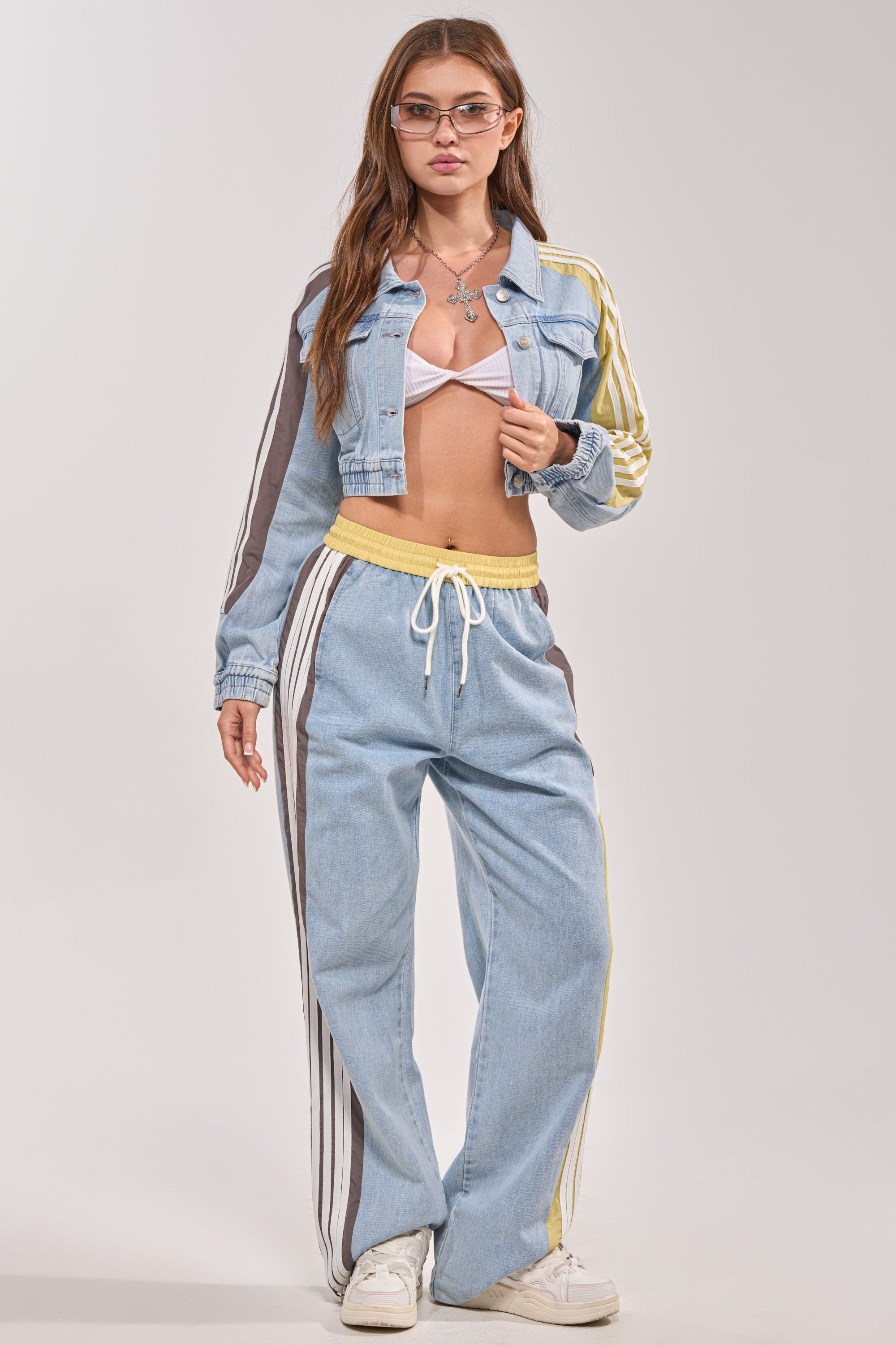 A woman in glasses wears the NEVER BETTER DENIM JACKET, featuring a cropped silhouette and striped sleeves, paired with a white bralette, light blue drawstring track pants with side stripes, and white sneakers against a plain background. 7