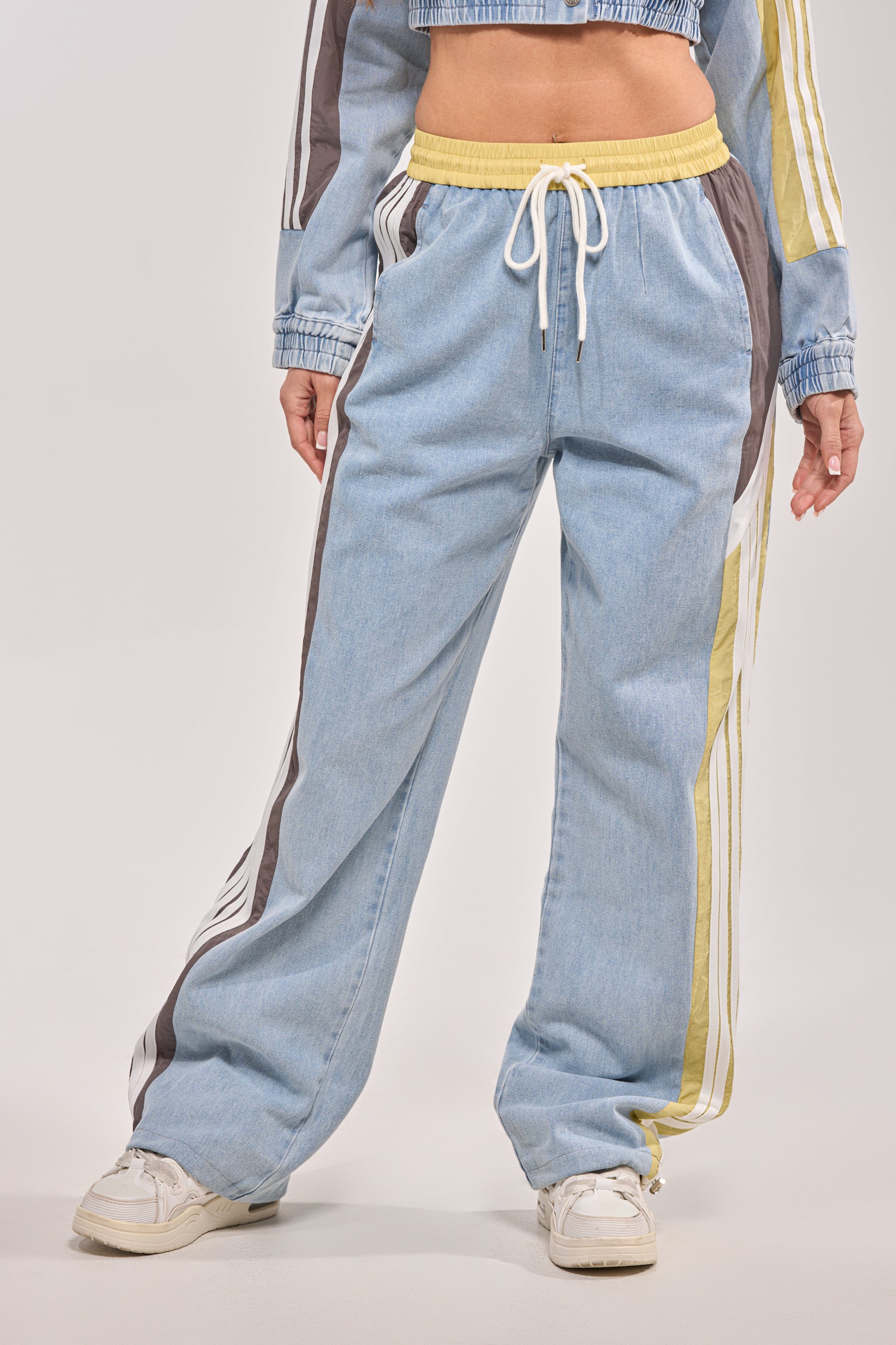 A person wears ALONE AGAIN DENIM TRACK PANT—light blue sporty pants with white, brown, and yellow side stripes, a high-rise elastic waistband—and white sneakers. Only the lower body and hands are visible.