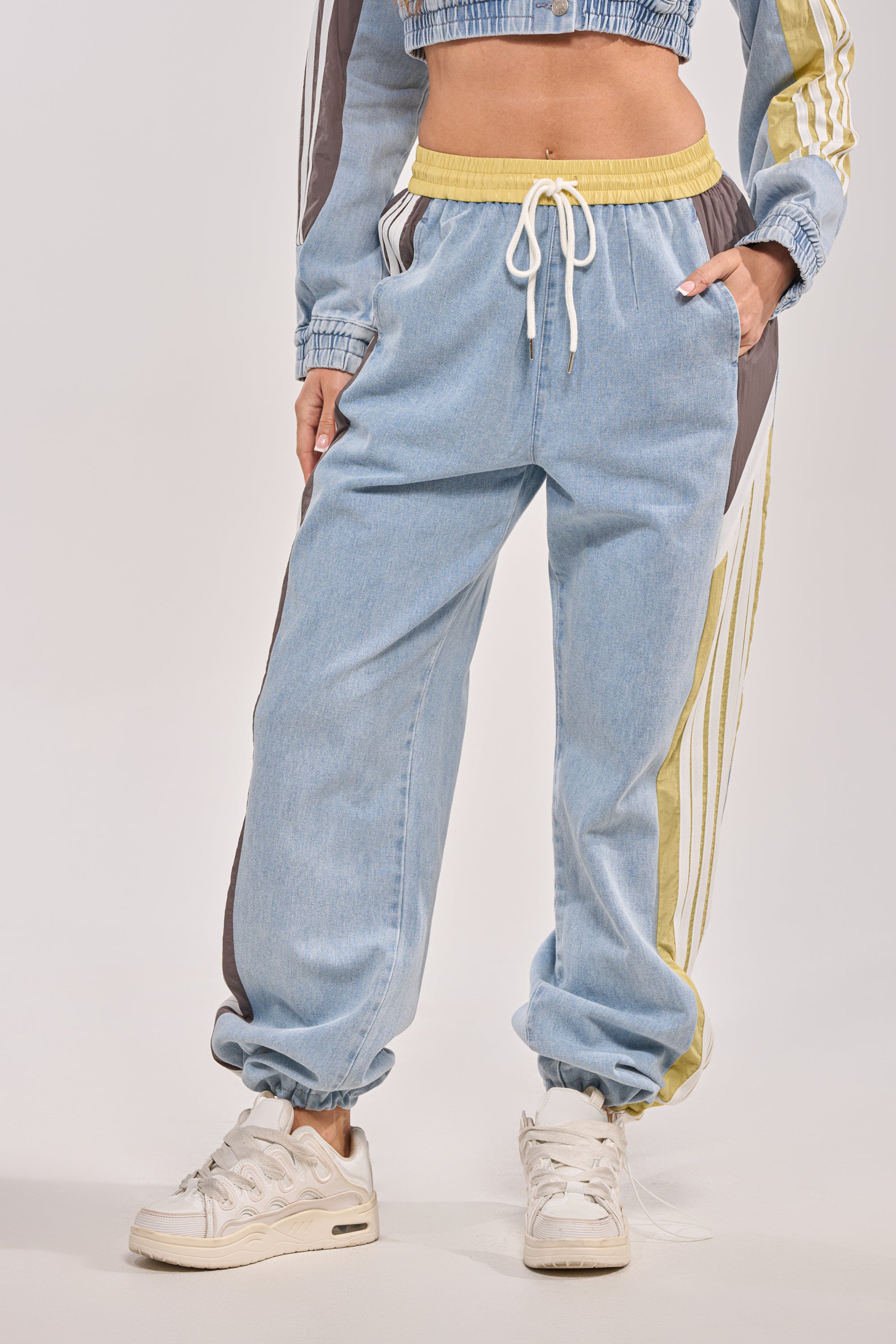 A person wears the ALONE AGAIN DENIM TRACK PANT, featuring light blue denim, a high-rise elastic waistband, and yellow and brown side stripes. The image focuses on the pants against a plain background.
