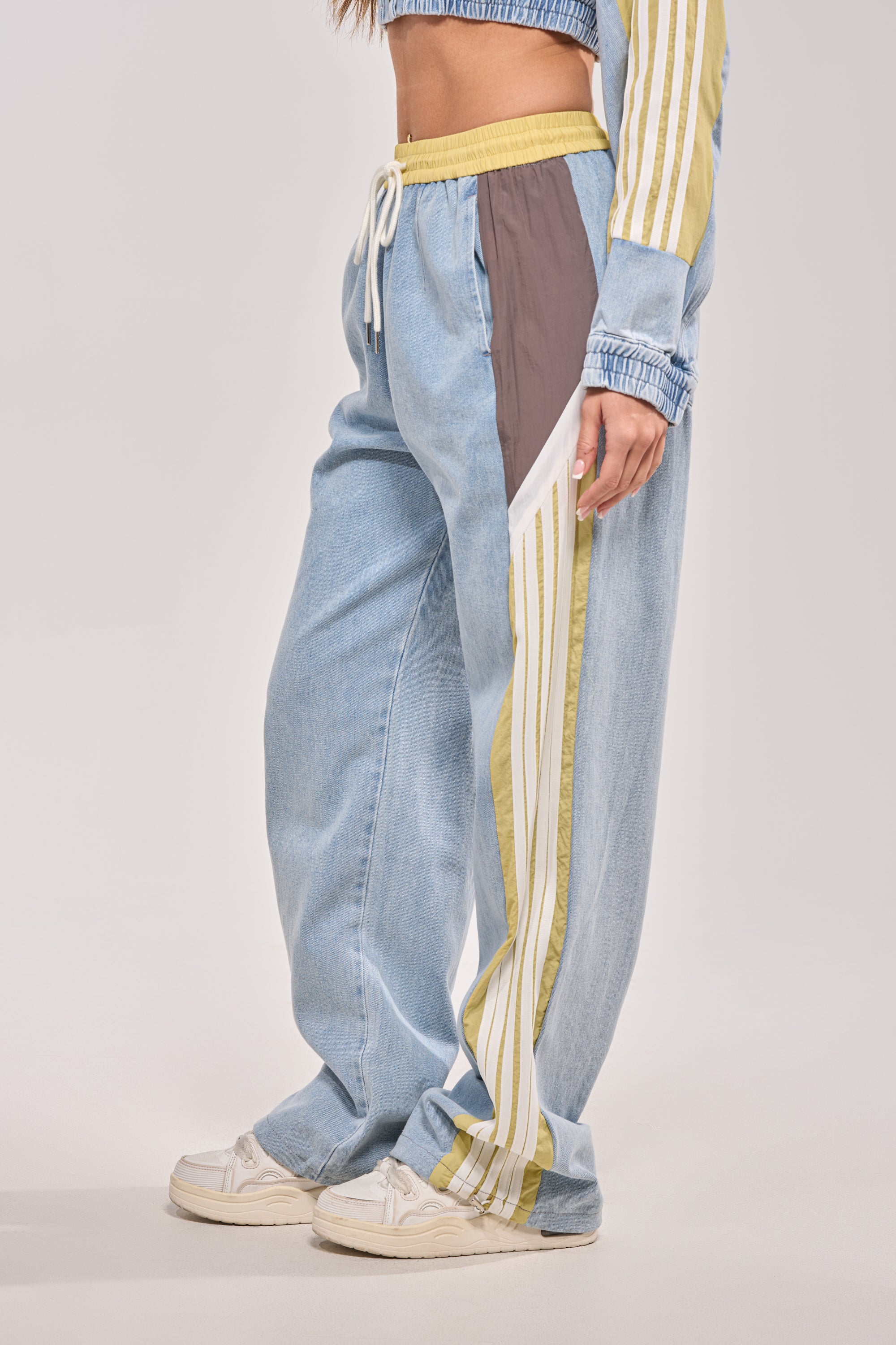 Wearing the ALONE AGAIN DENIM TRACK PANT in light blue with a high-rise elastic waistband and white and yellow side stripes, paired with white sneakers and a matching cropped jacket. The image is cropped at the shoulders.