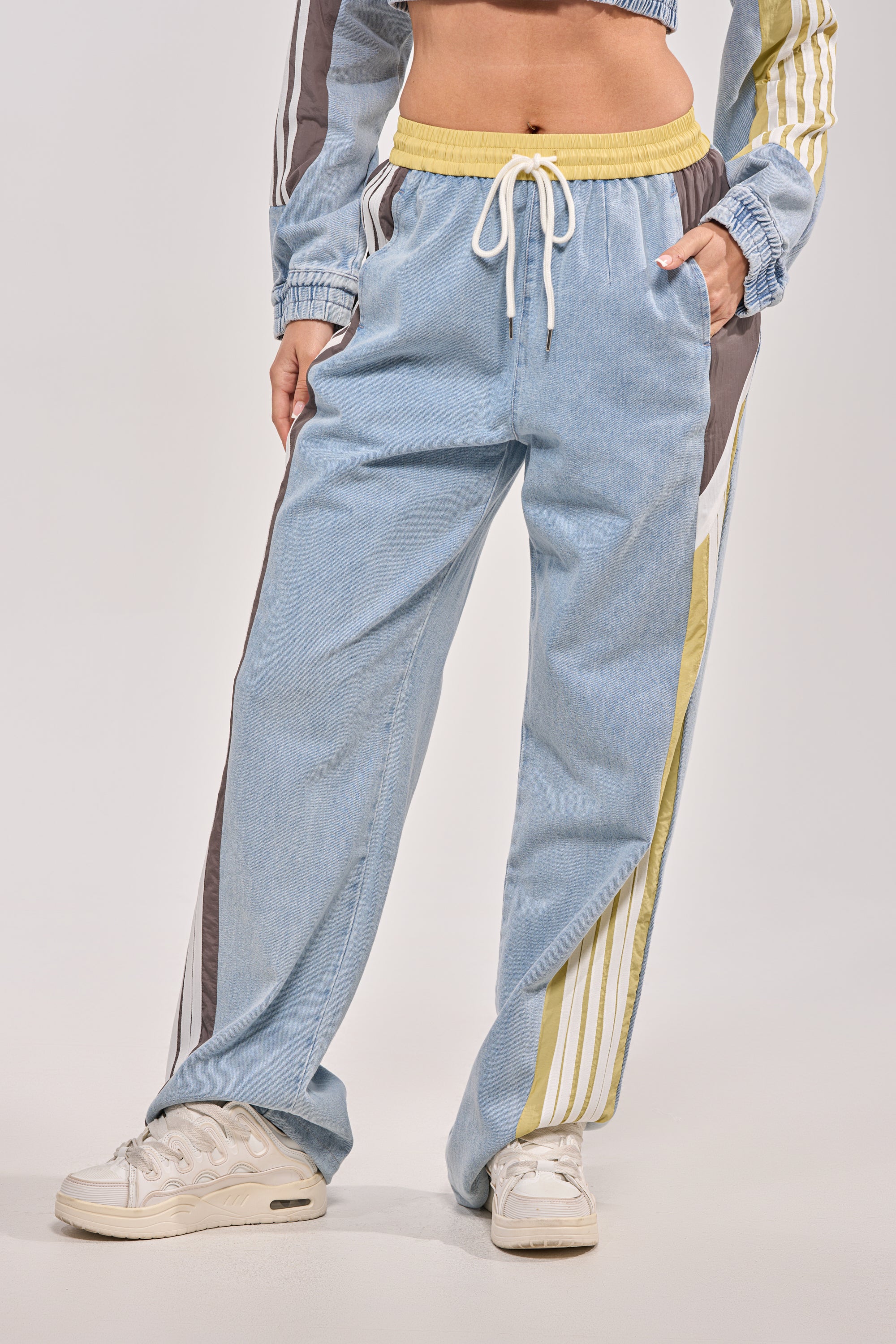A person wears ALONE AGAIN DENIM TRACK PANT—light blue joggers with yellow, brown, and white stripes, a high-rise elastic waistband with drawstrings—and white sneakers. The photo is cropped at the shoulders.