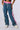 A person models the ALONE AGAIN DENIM PANT by GUANGZHOU EUROPE SOURCES, showcasing blue baggy jeans with white and pink striped inserts along the sides. The denim pants feature a contrast shirred elastic waistband and a white drawstring at the waist, accompanied by two side pockets. The person also wears white sneakers.