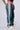 A person wearing the ALONE AGAIN DENIM PANT by GUANGZHOU EUROPE SOURCES, which features blue baggy jeans with vertical white and gray stripes on the sides, a pink contrast shirred elastic waistband, and white drawstrings. They are also sporting white sneakers. The wide-leg silhouette adds an extra layer of flair as their hands rest in their pockets, with the upper body cropped out.