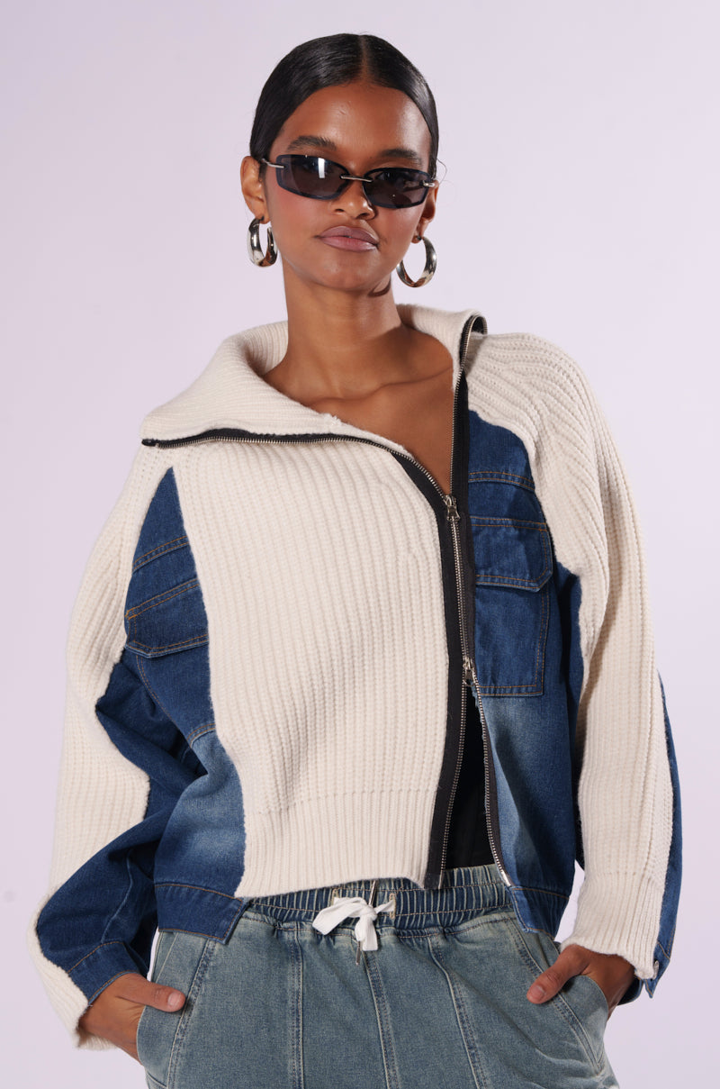 A person with dark hair tied back, wearing oval sunglasses and large hoop earrings, confidently poses. They are stylishly dressed in the SOMETHING ABOUT YOU SWEATER from the AKIRA Label, which features denim paneling, paired with drawstring waist jeans. Hands are casually placed in their pant pockets.