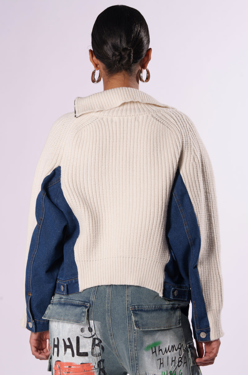 A person with dark, tied-back hair is seen from behind wearing the SOMETHING ABOUT YOU SWEATER, a chunky white knitted piece accented with blue denim patches on its woven knit base. They also sport light blue jeans decorated with abstract, colorful designs and text. Large hoop earrings complete their look against a plain, light-colored background.