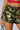 A person is shown from the waist down, wearing the UP ALL NIGHT CAMO SHORT with various patches and embellishments, including a "WHO CARES" patch. They have a bejeweled belly button piercing and are holding a striped handle bag. The person is wearing a red top.