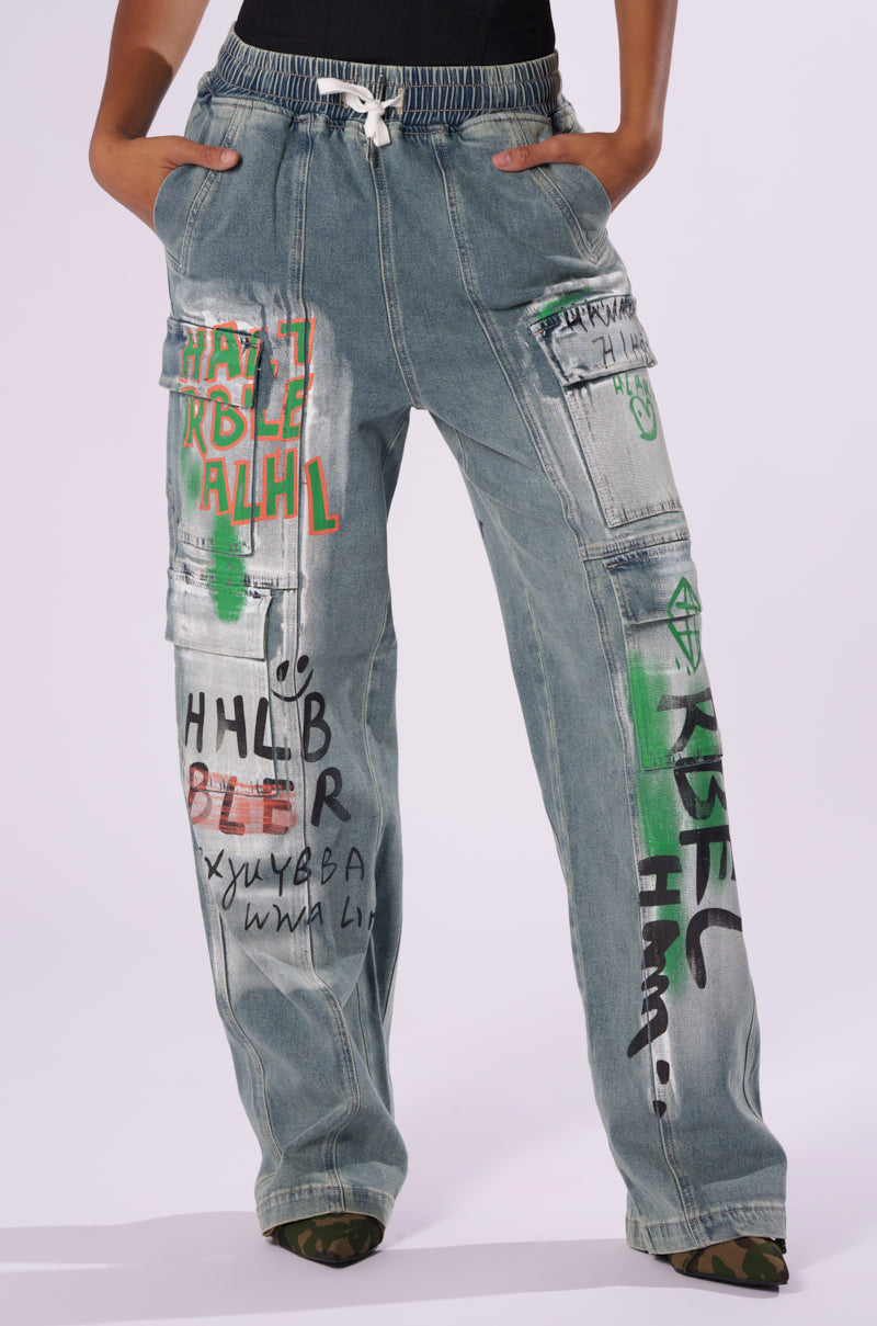 A person wearing the "CHANGES EVERYTIME DENIM PANT" in light blue, featuring a high-rise and wide-leg silhouette. The jeans are adorned with colorful graffiti-inspired graphics, including green and red phrases, abstract shapes, and a mix of symbols. They also have a drawstring waist and side pockets. The background is plain white.