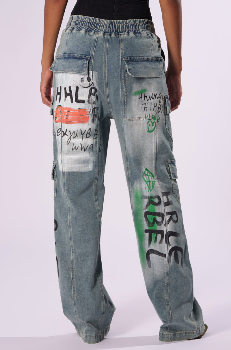 A person wearing the CHANGES EVERYTIME DENIM PANT, which features loose-fitting, high rise denim with hand-drawn graffiti-inspired graphics in various colors on the back. These jeans include multiple pockets and a gathered waistband, showcasing a wide leg silhouette against a plain white background.