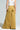 Rear view of a person wearing a white tank top and the GIVE IT TO ME LOW RISE DENIM PANT, featuring wide-legged, mustard-colored fabric with large back pockets. The photo highlights the flared silhouette of the pants' design.