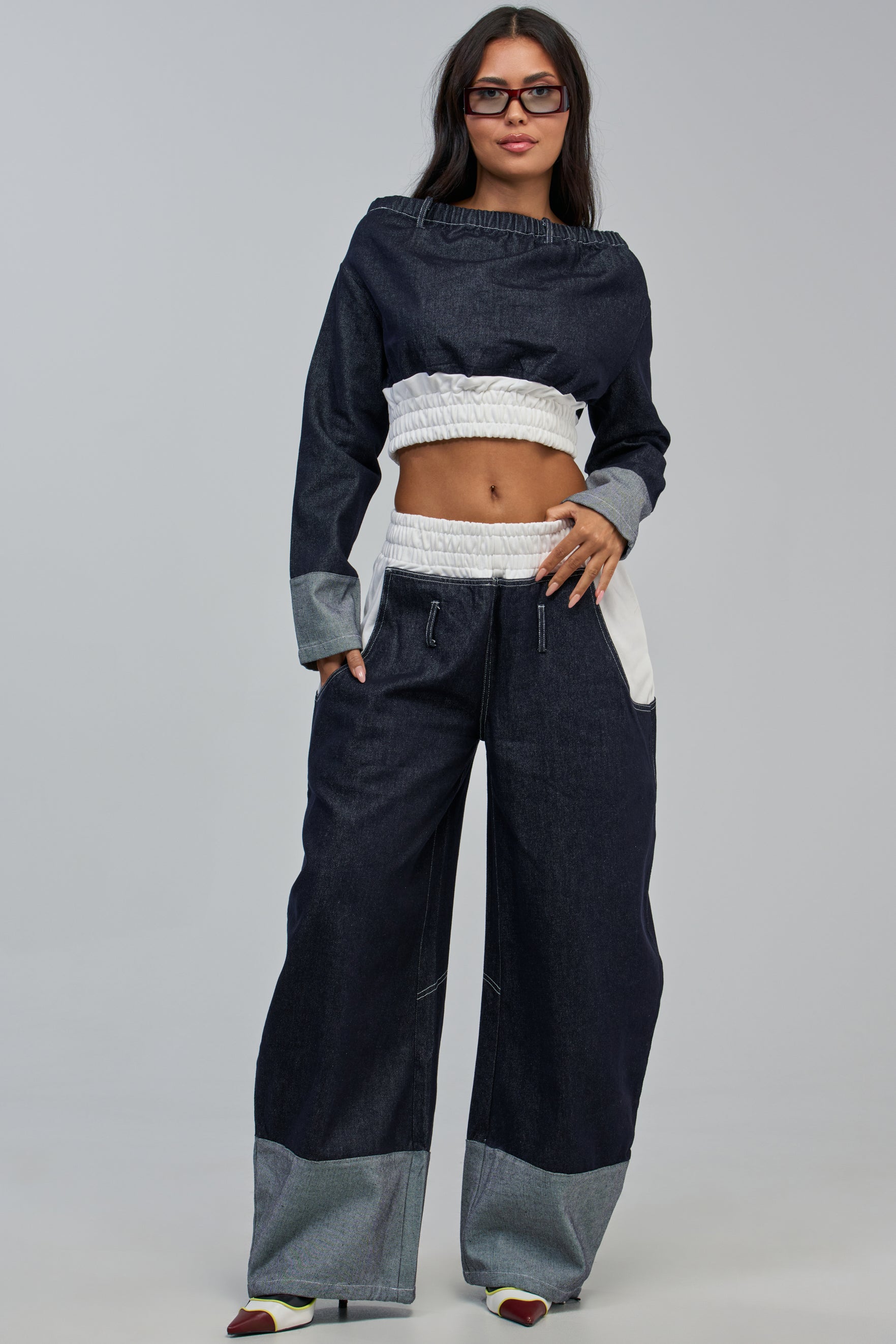 Model showcasing the WHO SHE WIDE LEG JOGGER in dark denim paired with a matching crop top featuring light gray cuffs and white accents. She stands confidently against a plain background, wearing glasses and exuding an effortlessly cool streetwear vibe.