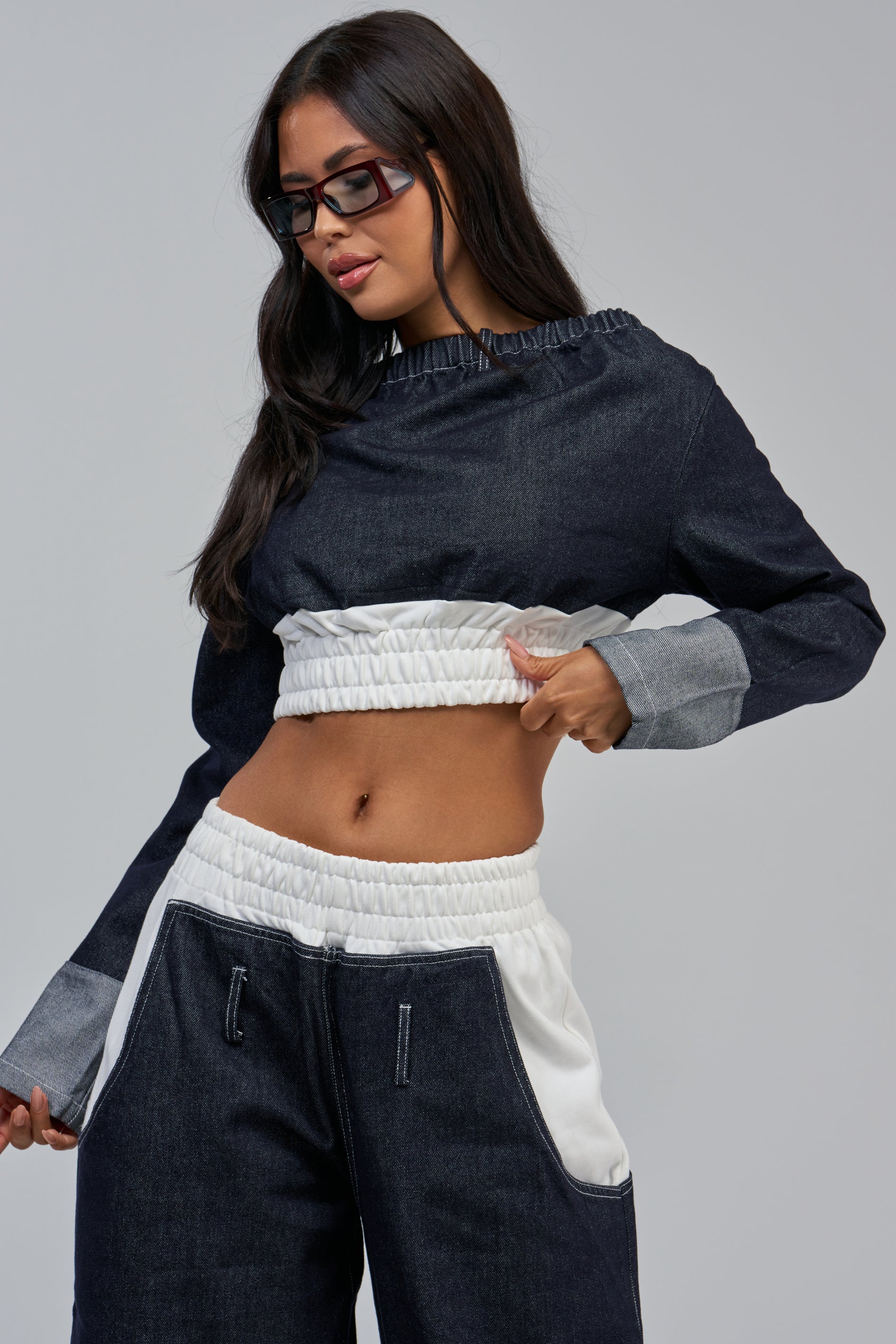 Wearing the WHO SHE OFF THE SHOULDER DENIM SWEATSHIRT paired with matching denim pants featuring white accents, a woman in sunglasses channels streetwear vibes as she poses against a plain background.