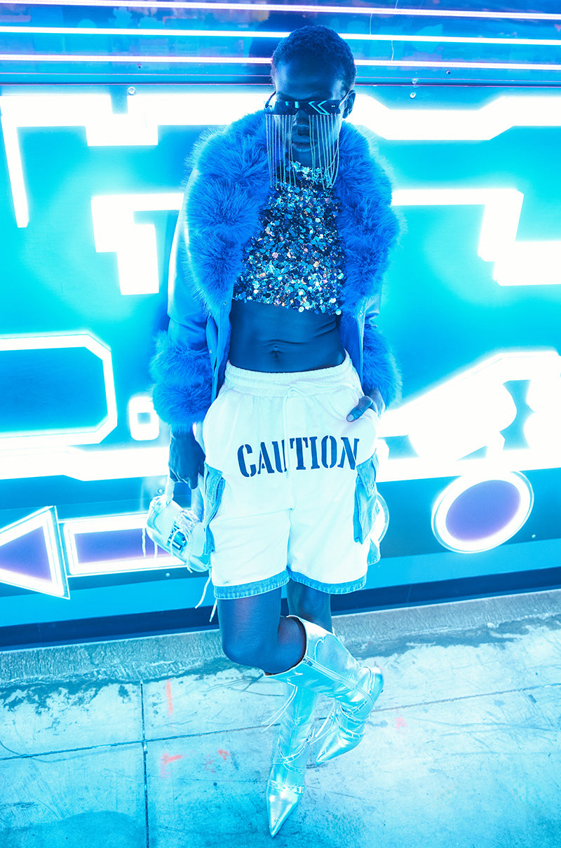 Beneath the neon blue lights, a fashion model strikes a pose in a glittery top and fluffy blue coat. The PROCEED WITH CAUTION SHORT, featuring edgy denim cargo pockets, is stylishly combined with silver high boots to create a futuristic vibe that amplifies the vibrant ensemble.