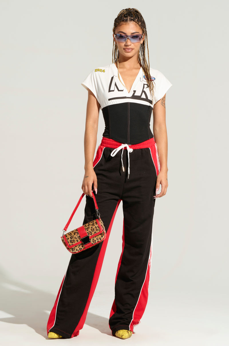 A person with long braided hair wearing dark sunglasses, a white graphic t-shirt, and a pair of WE GOTTA GO PANT - black and red athletic pants with a wide-leg silhouette and white stripes - holds a leopard print handbag. They stand confidently against a neutral background.