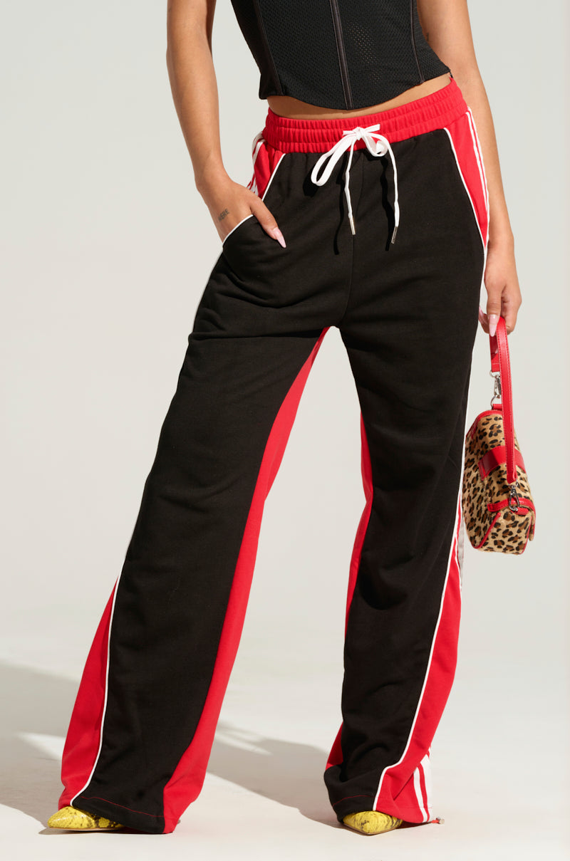 A person sporting the WE GOTTA GO PANT, a chic pair of black and red track pants featuring white drawstrings and side stripes, along with a colorblock pattern. They pair it with a sleek black top and hold a small leopard print bag that has a striking red handle. The image captures only the lower half of their body against an unadorned backdrop.