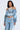 A person wears an off-the-shoulder LEILA Denim Patchwork Sweatshirt and light-wash jeans, accessorized with a statement pearl necklace and hoop earrings against a light gray background.