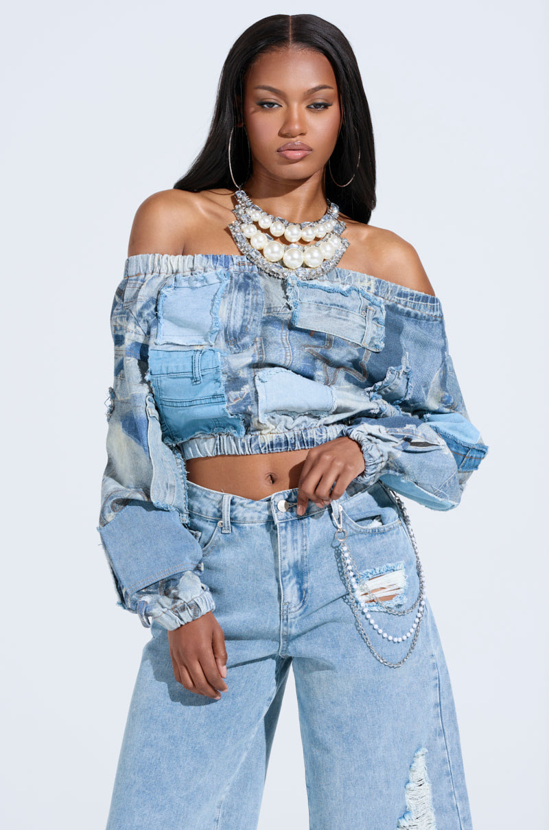 A woman wears a LEILA Denim Patchwork Sweatshirt off-shoulder style paired with distressed jeans. Her look is highlighted by large hoop earrings, a layered pearl necklace, and her long straight hair flowing elegantly against a plain background.