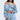 A woman wears a LEILA Denim Patchwork Sweatshirt off-shoulder style paired with distressed jeans. Her look is highlighted by large hoop earrings, a layered pearl necklace, and her long straight hair flowing elegantly against a plain background.