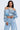 A woman wears a LEILA Denim Patchwork Sweatshirt off-shoulder style paired with distressed jeans. Her look is highlighted by large hoop earrings, a layered pearl necklace, and her long straight hair flowing elegantly against a plain background.