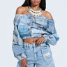 A woman wears a LEILA Denim Patchwork Sweatshirt off-shoulder style paired with distressed jeans. Her look is highlighted by large hoop earrings, a layered pearl necklace, and her long straight hair flowing elegantly against a plain background.