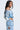 A woman with long hair models the LEILA Denim Patchwork Sweatshirt, pairing it with high-waisted jeans and a pearl necklace. She stands against a plain background, gazing to her right.