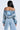 A long-haired person faces away, showcasing the silhouette of a LEILA Denim Patchwork Sweatshirt that's off-the-shoulder, paired with light blue jeans. A small tattoo graces their right shoulder against a plain, light backdrop.