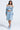 A woman strides confidently in the LEILA Denim Patchwork Sweatshirt, styled off-the-shoulder with a cropped silhouette. She pairs it with a distressed knee-length skirt, chunky necklace, and fluffy heels against a plain, light background.
