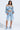 A woman confidently wears the LEILA DENIM PATCHWORK SWEATSHIRT, styled as a light blue off-the-shoulder top with knee-length ripped shorts. Layered necklaces and fluffy blue heeled shoes complete her chic ensemble against a plain background.