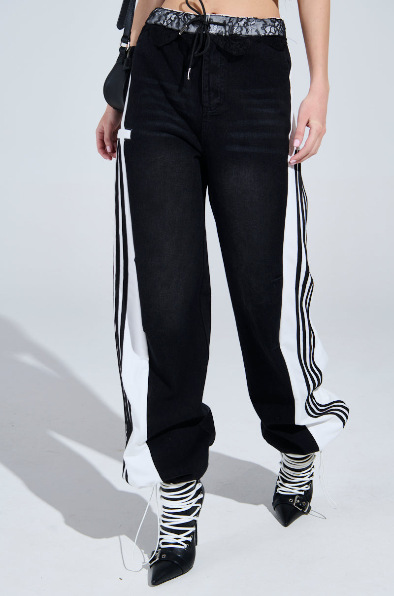 Wearing SPORTY AND SWEET BLACK WASH DENIM TRACK PANTS with white vertical stripes for a sporty-chic look, the ensemble includes black pointed high-heels with white laces and a sleek black bag, all set against a plain light gray background.