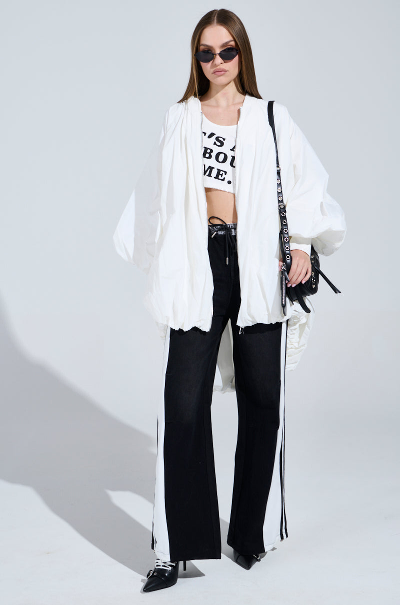 A woman exudes a sporty-chic vibe, confidently posing in high-rise SPORTY AND SWEET BLACK WASH DENIM TRACK PANTS, paired with a white oversized jacket, cropped graphic shirt, pointed shoes, and a black handbag against a plain background.