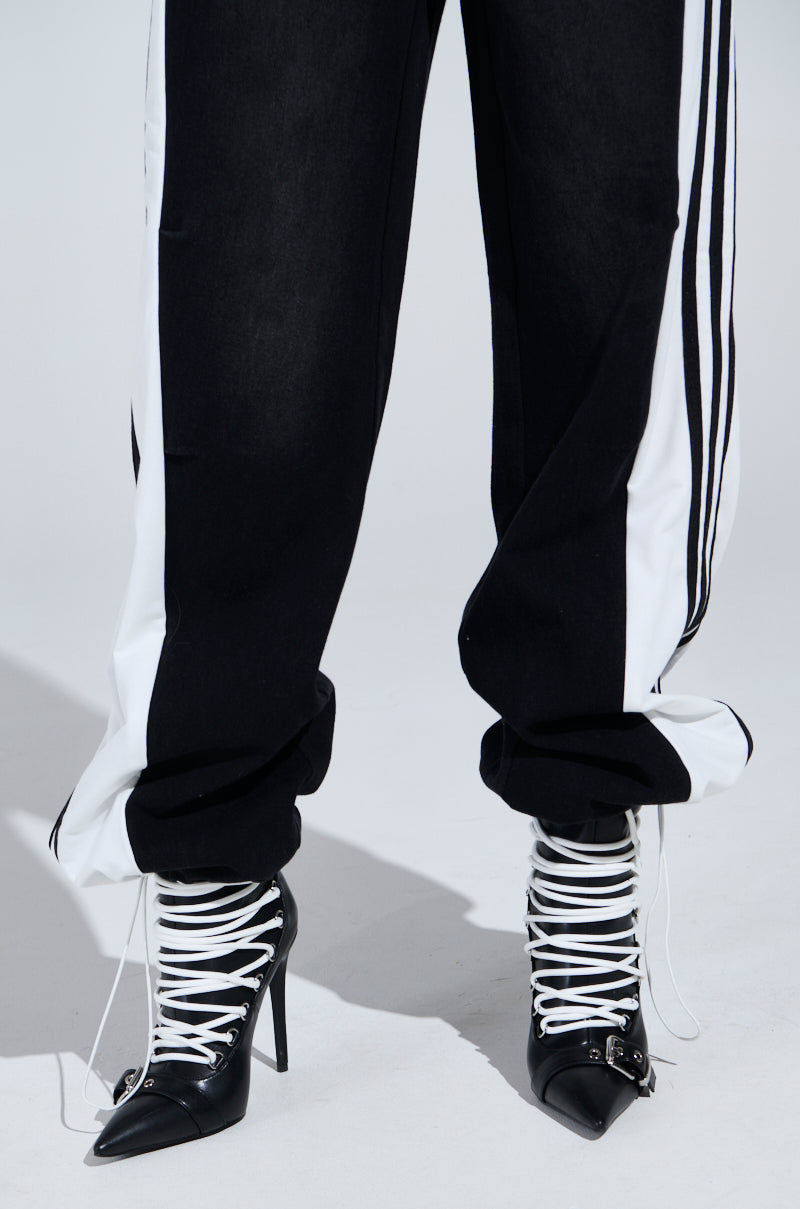 Dressed in high-rise black SPORTY AND SWEET DENIM TRACK PANTS with white side stripes, paired with black high-heeled boots featuring crisscrossing white laces. The slightly scrunched pants add a relaxed flair against the plain, light background.