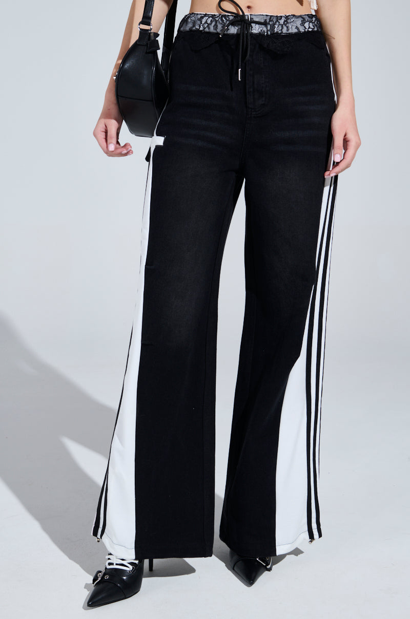 The lower half of a person in black and white Sporty and Sweet Black Wash Denim Track Pants with wide legs and side stripes is visible. They hold a black handbag and wear black pointed shoes against a neutral background.