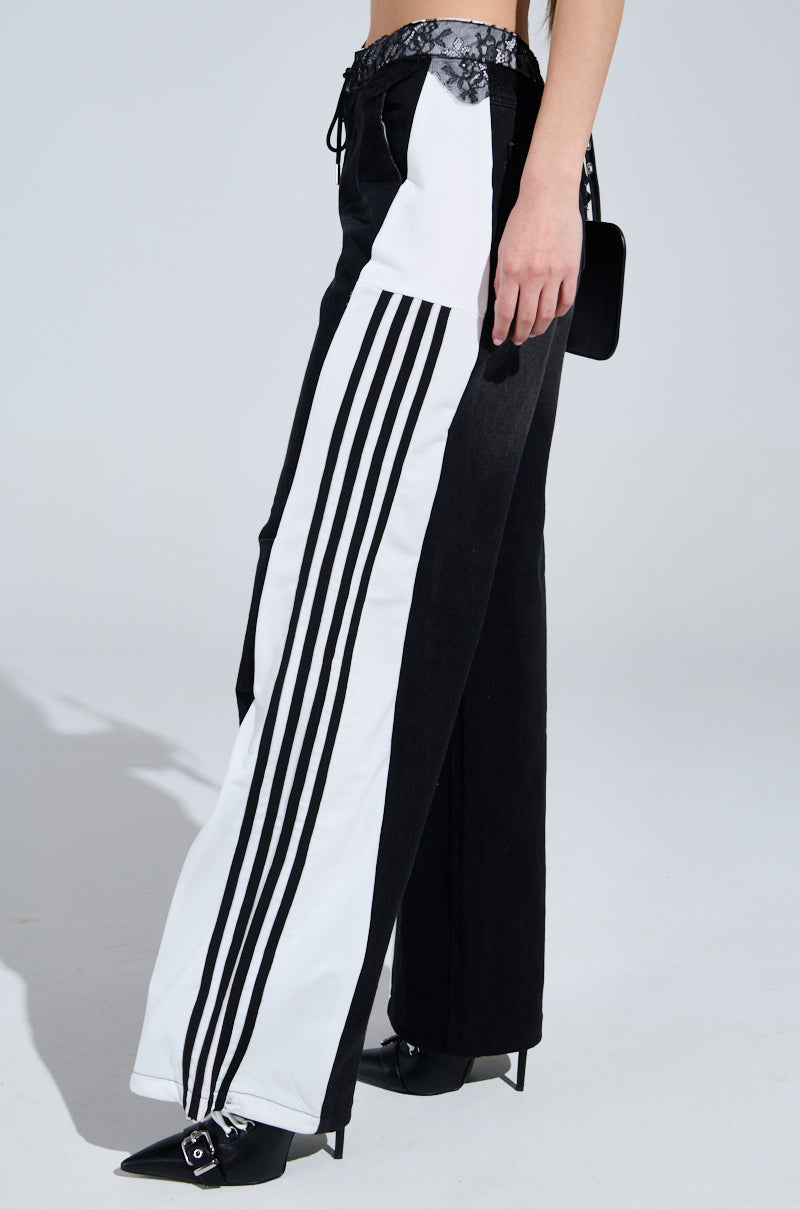 Dressed in sporty-chic SPORTY AND SWEET BLACK WASH DENIM TRACK PANTS with side stripes, paired with pointed black shoes and a small black bag, the person stands against a plain, light-colored backdrop.