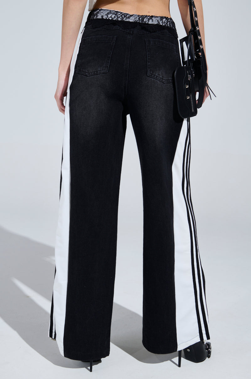 A person is wearing Sporty and Sweet Black Wash Denim Track Pants, featuring chic black denim with white side panels and black stripes. They're accessorized with a lace waistband, a black shoulder bag, and paired with sleek black pointed shoes.