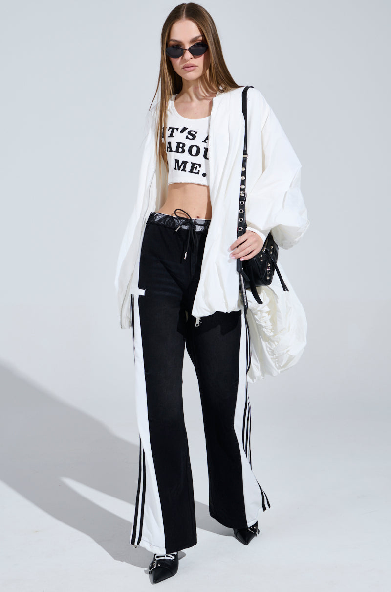 A woman exudes sporty-chic confidence in a white crop top under a matching oversized jacket, paired with SPORTY AND SWEET BLACK WASH DENIM TRACK PANTS and black heels. Her black purse complements the look as she stands poised against a light grey background.