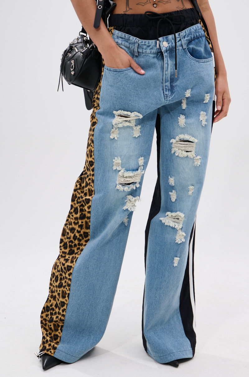 A person wearing WILDIN' OUT DENIM TRACKPANTS with leopard-print panels, black stripes, and frayed holes, paired with a black crossbody bag, stands against a plain white background. 0
