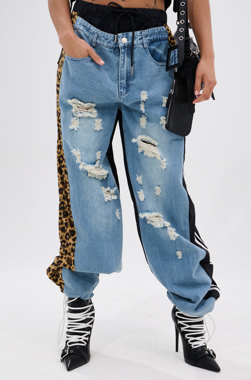 WILDIN' OUT DENIM TRACKPANTS featuring distressed blue denim with leopard-print panels are styled with a black belt, lace-up heeled boots, and a black purse against a plain white background. 1