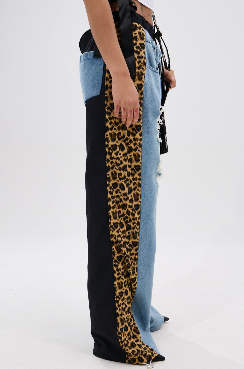 Model wears the WILDIN' OUT DENIM TRACKPANTS—high-waisted, distressed jeans with black side panels and a vertical leopard-print stripe—posed against a plain white background. 2