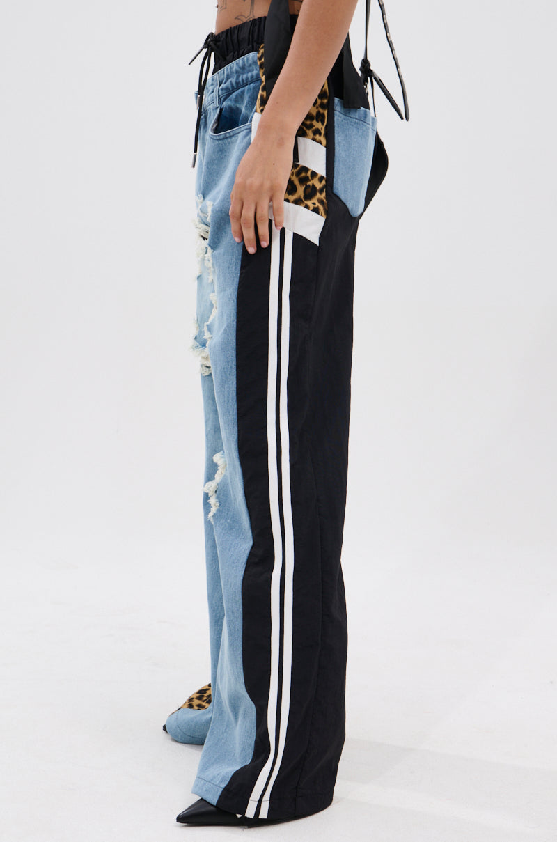 A person wears WILDIN' OUT DENIM TRACKPANTS with a half-denim, half-black design, white side stripes, distressed details, and carries a leopard print bag. The side-view photo is set against a plain background. 3