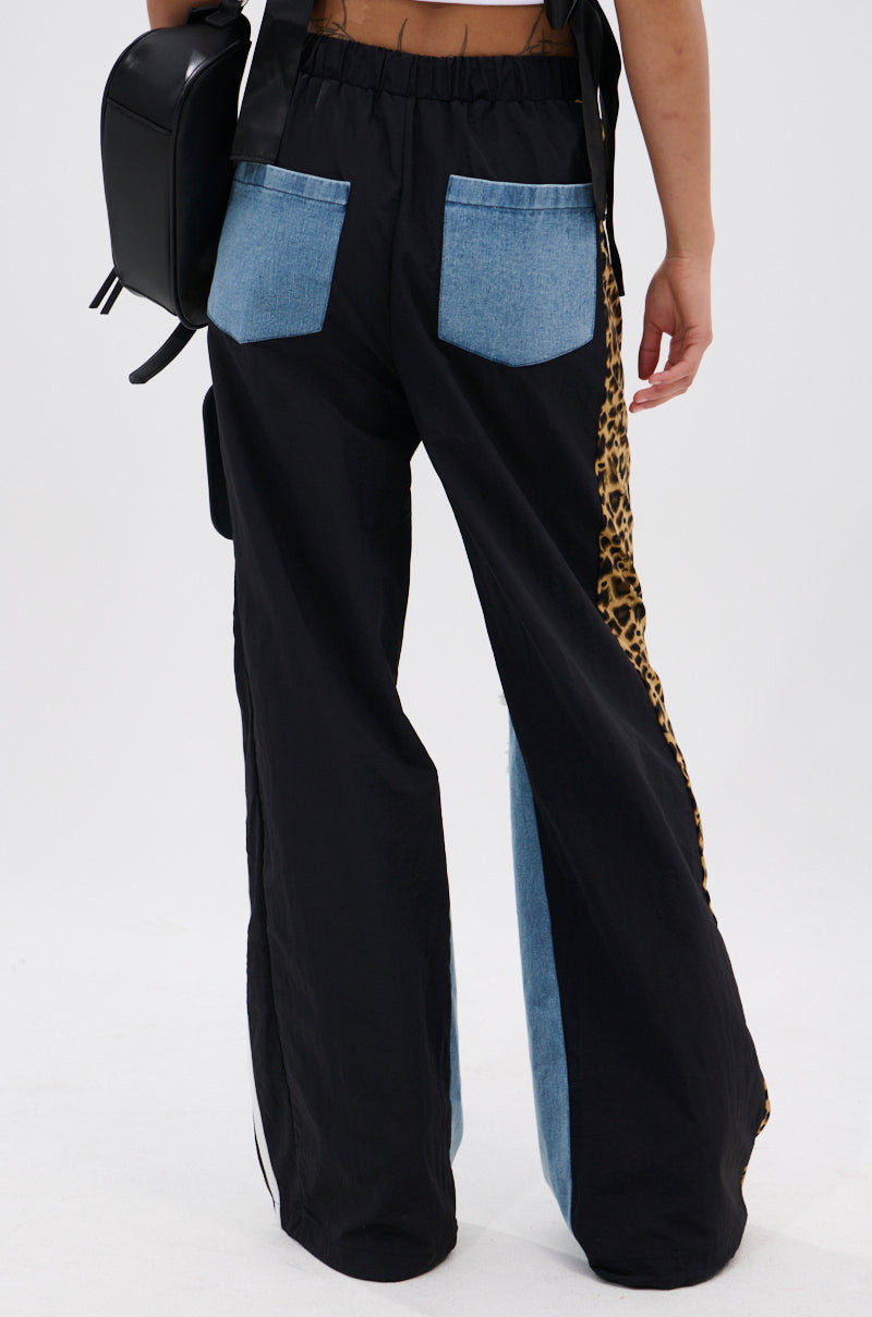 A person models the WILDIN' OUT DENIM TRACKPANTS, featuring wide legs, blue denim back pockets, and leopard-print side panels. They hold a black bag and stand against a plain white background. 4