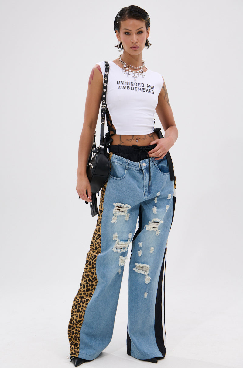A woman stands confidently in a white "UNHINGED AND UNBOTHERED" crop top, WILDIN' OUT DENIM TRACKPANTS with leopard-print panels, a black belt, layered necklaces, hoop earrings, and a black crossbody bag. 5