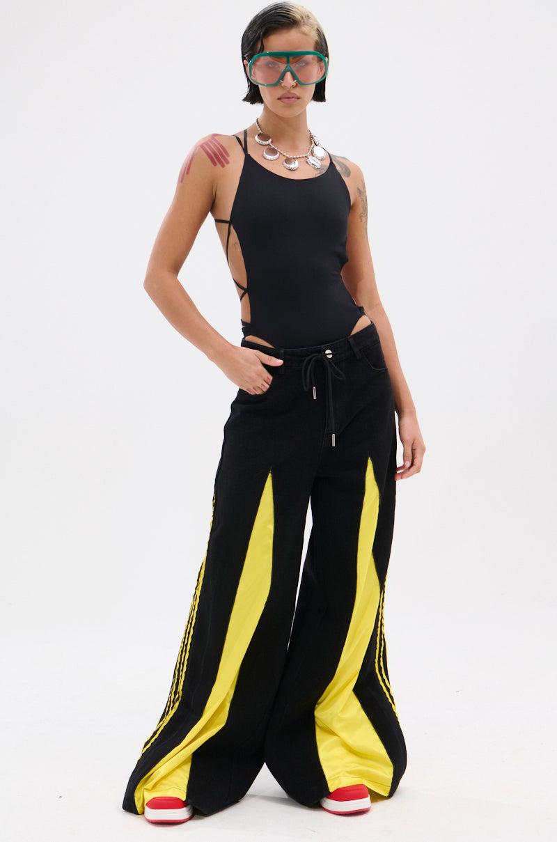 A person poses confidently against a white background wearing the GAME TIME DECISION DENIM PANT, featuring wide legs with bold yellow panels, paired with a black cutout bodysuit, red-and-white sneakers, chunky necklace, and large green sunglasses.