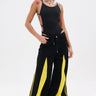 A person poses confidently against a white background wearing the GAME TIME DECISION DENIM PANT, featuring wide legs with bold yellow panels, paired with a black cutout bodysuit, red-and-white sneakers, chunky necklace, and large green sunglasses.