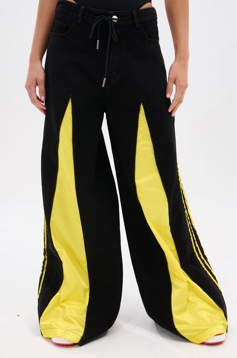A person wears the GAME TIME DECISION DENIM PANT—wide-leg black jeans with bold yellow panels down the front and sides, featuring a drawstring waist. Hands rest at their sides against a plain, light background.